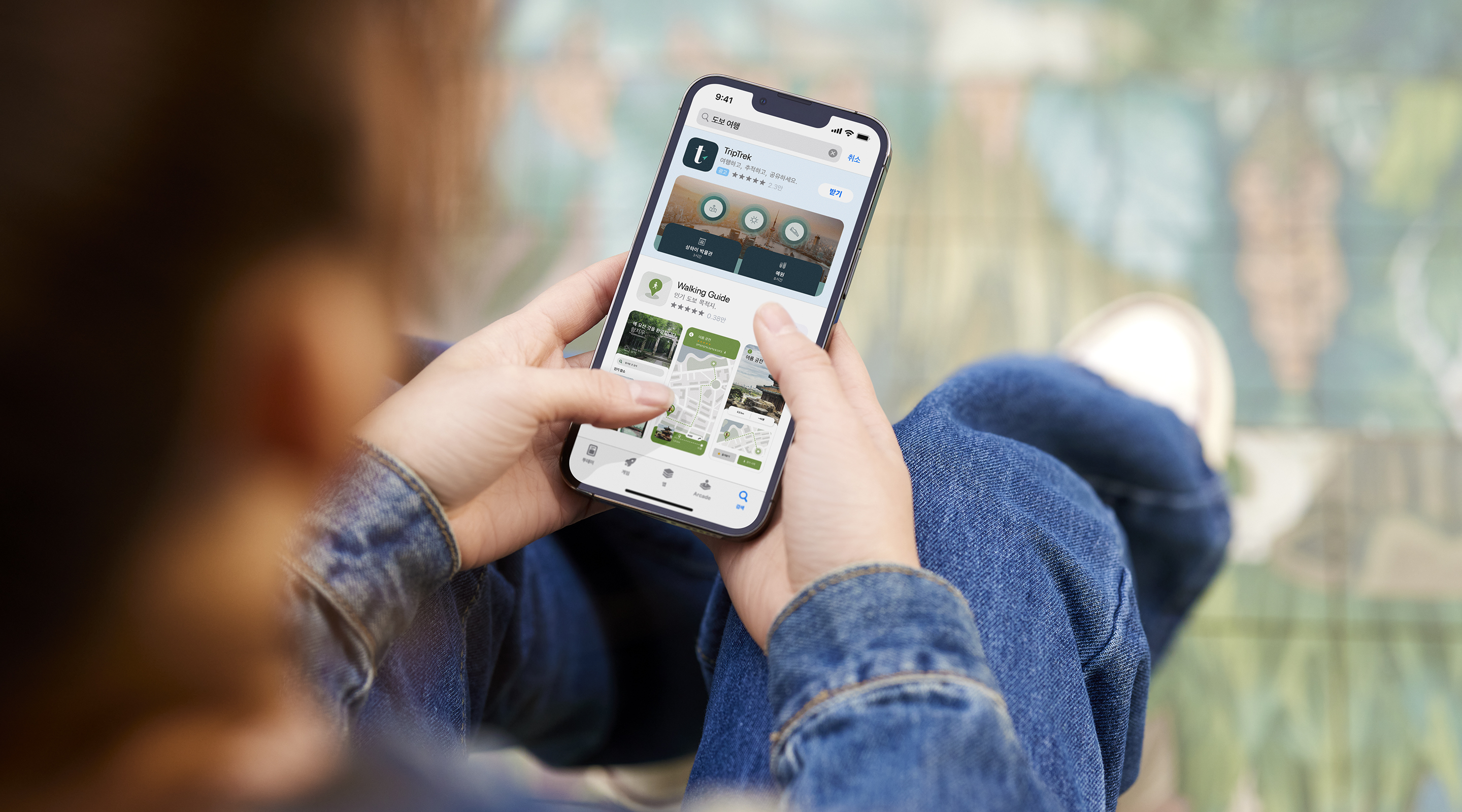 양손으로 iPhone을 들고 있습니다. App Store에 예제 앱 TripTrek의 검색 결과 광고가 열려 있고 검색 상자에 검색어 ‘걷기 여행’이 입력되어 있습니다. 