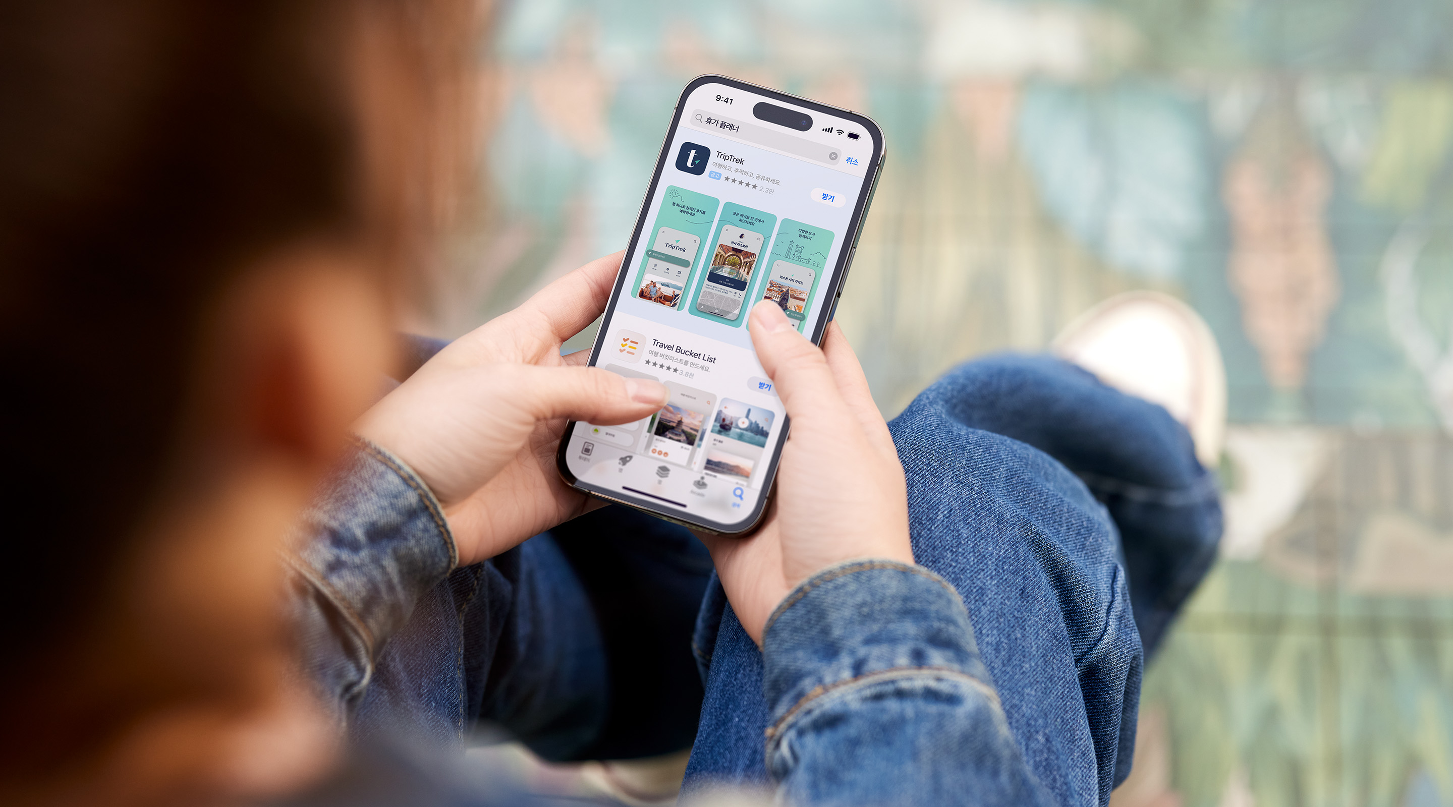 양손으로 iPhone을 들고 있습니다. App Store에 예제 앱 TripTrek에 대한 검색 결과 광고가 표시되어 있고, 검색 상자에 검색어 '휴가 플래너'가 입력되어 있습니다.