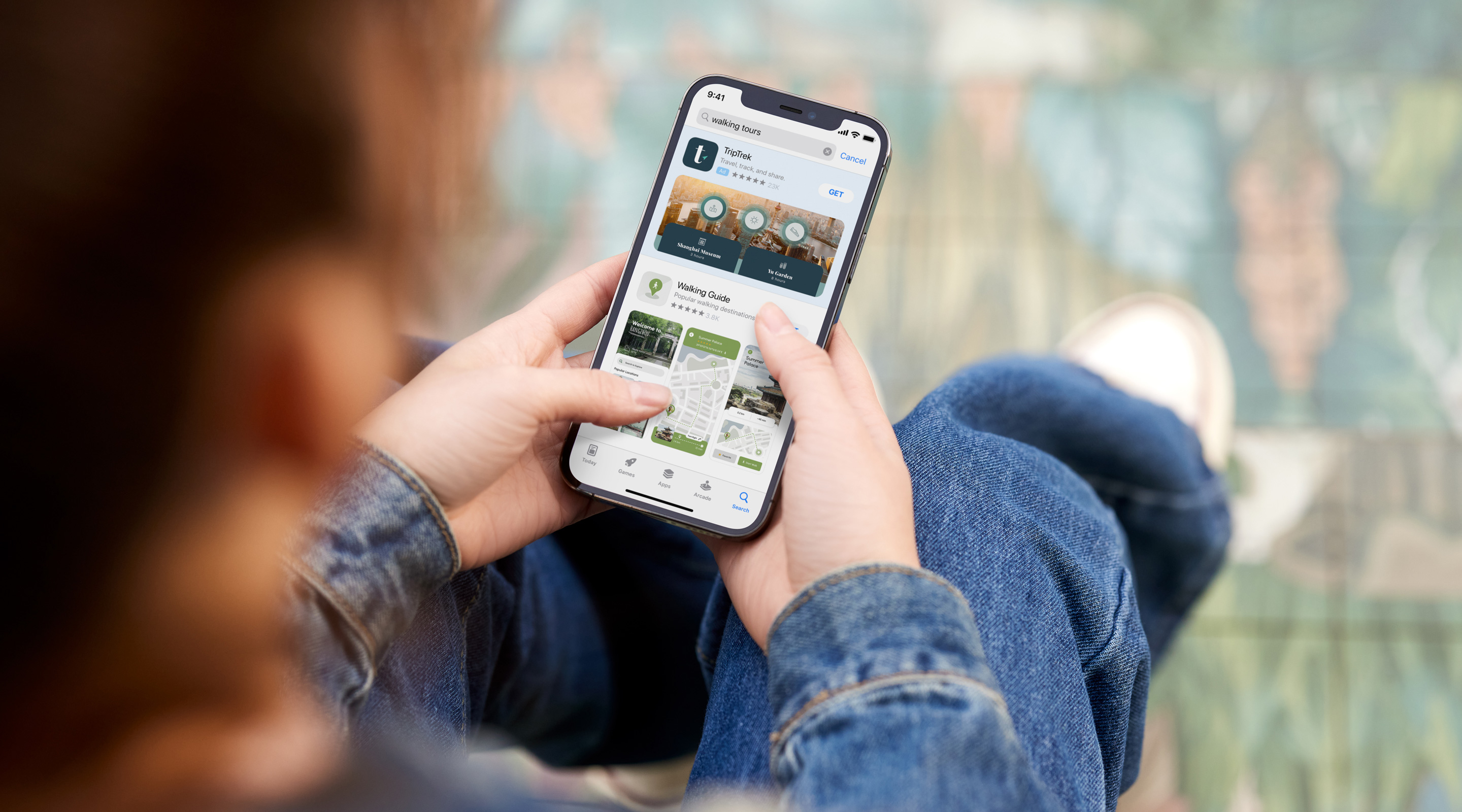 Two hands holding an iPhone. The App Store is open to a search results ad for the example app, TripTrek, with the term "walking tours" entered in the search box. 