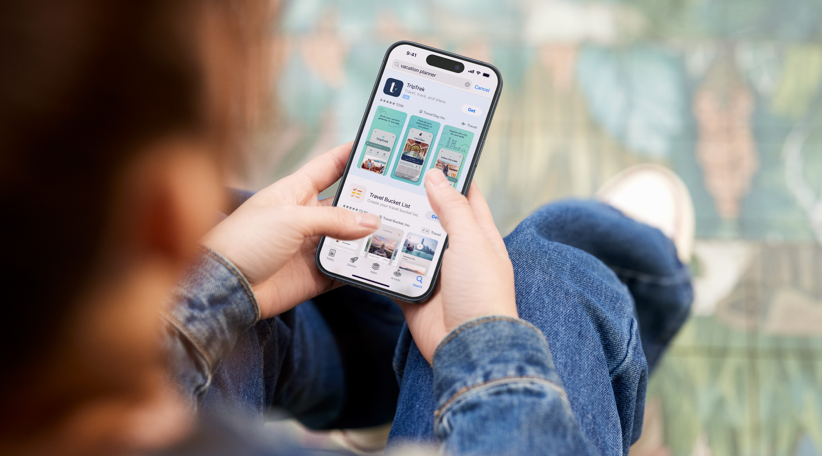 Two hands holding an iPhone. The App Store is open to a search results ad for the example app, TripTrek, with the term "vacation planner" entered in the search box. 